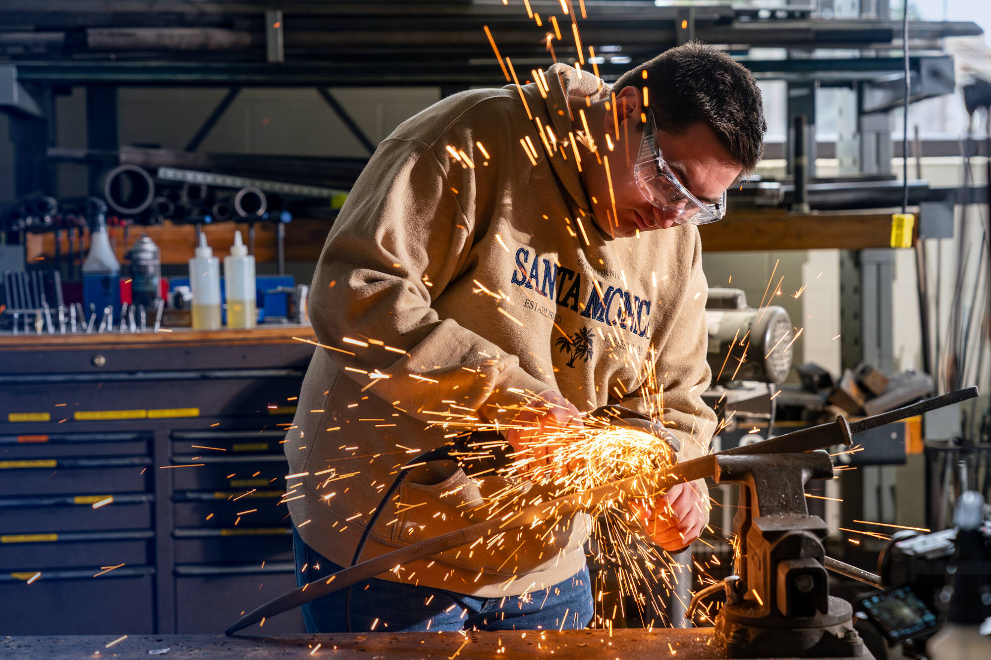 An engineering student wearing protective goggles sends sparks flying as they use a grinder on a piece of metal in a workshop.